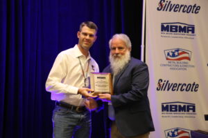 A man with a gray beard presenting Travis Wendt, with Metallic Products, with the MBMA Safety Award on a stage in front of a blue background